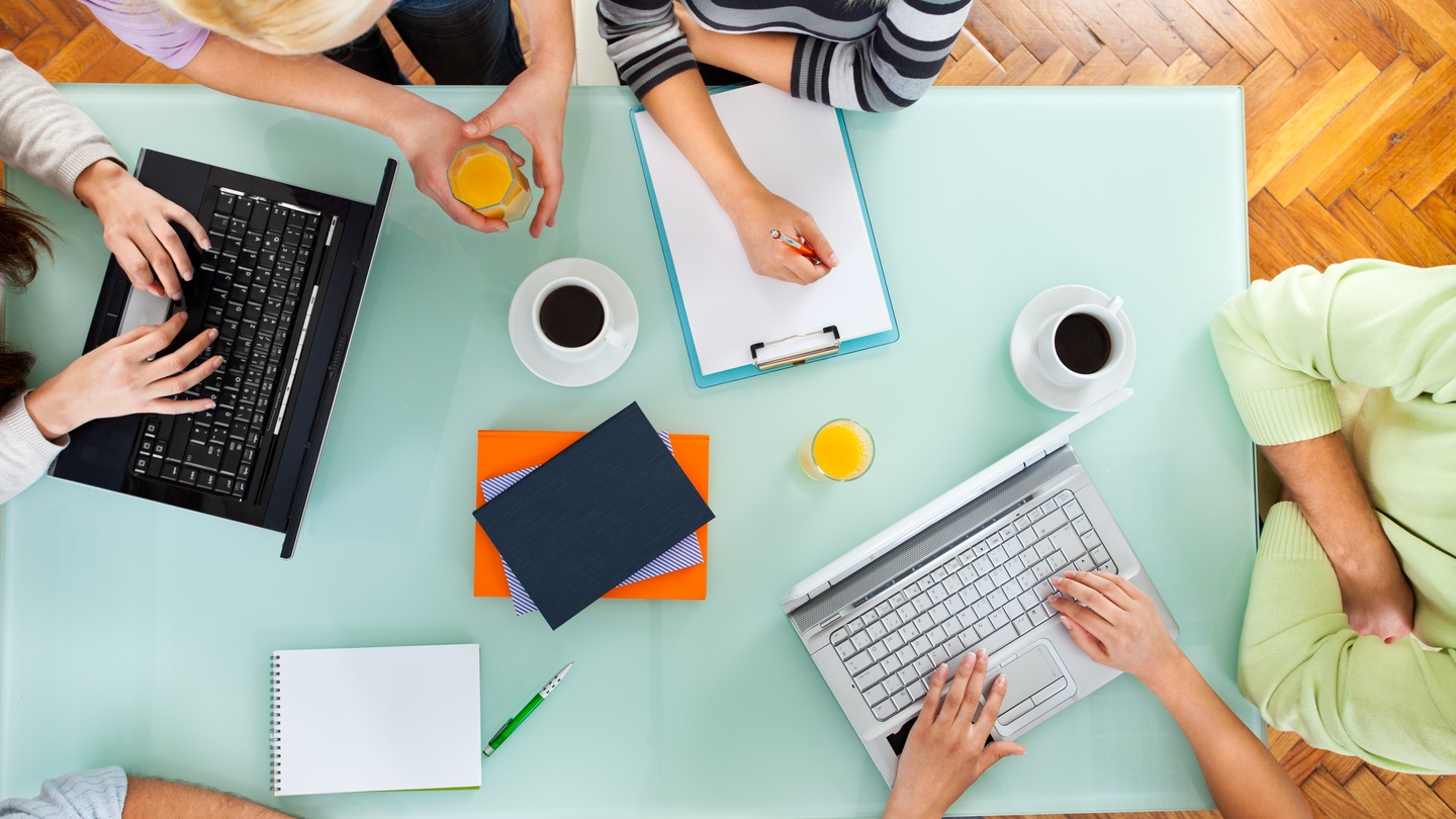 Several people sit at a table to write together, some are using laptops while others are using pens and paper
